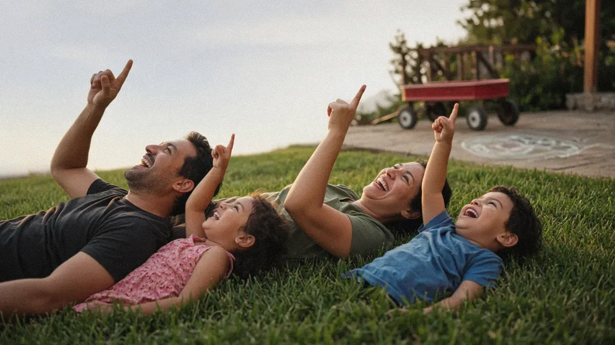 A mother, father, and their two young children lie on the backyard grass at golden hour, laughing and pointing at the sky